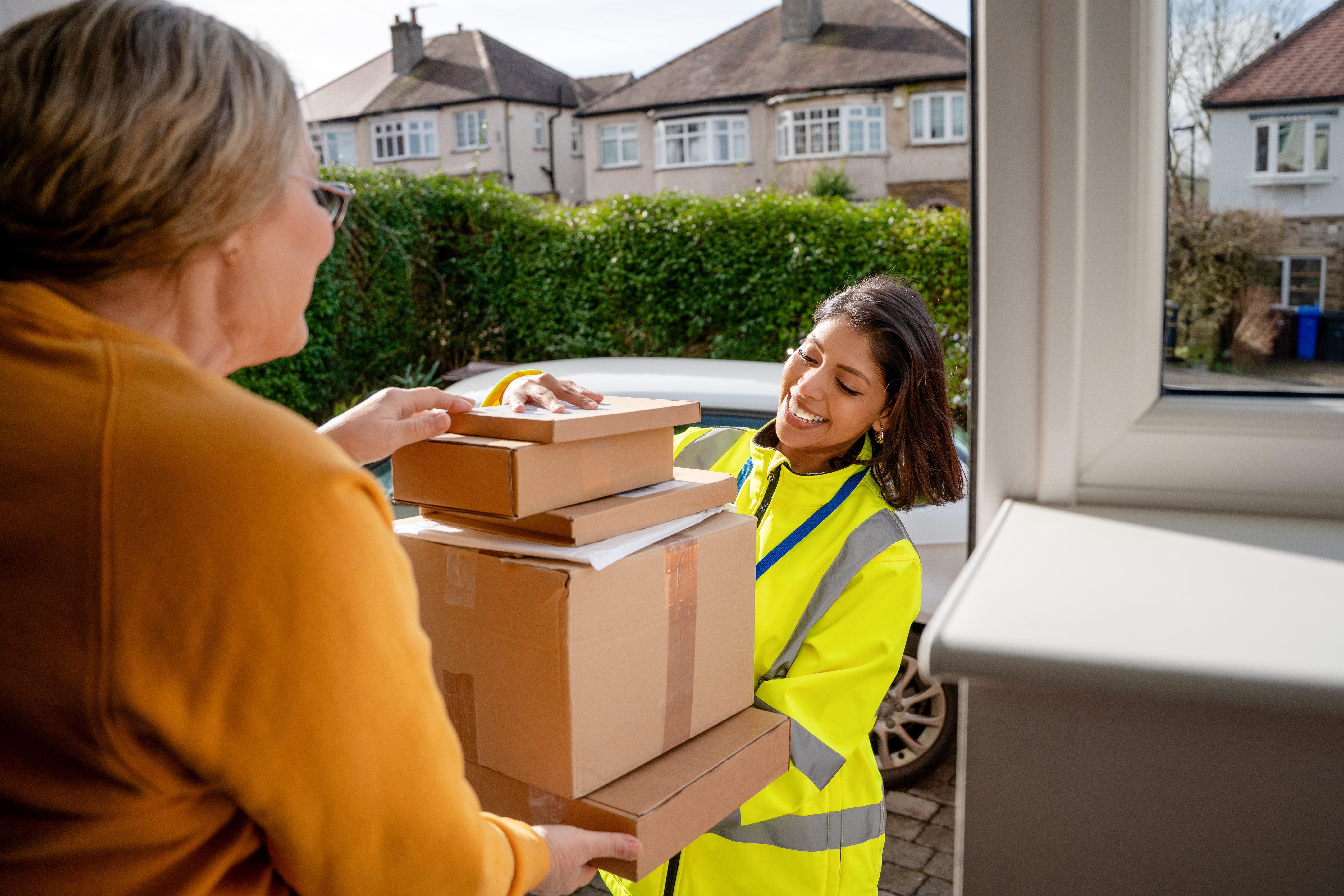 A delivery worker brings multiple packages to a woman at her house. The worker smiles while handing over the boxes. It is daytime in a neighborhood.